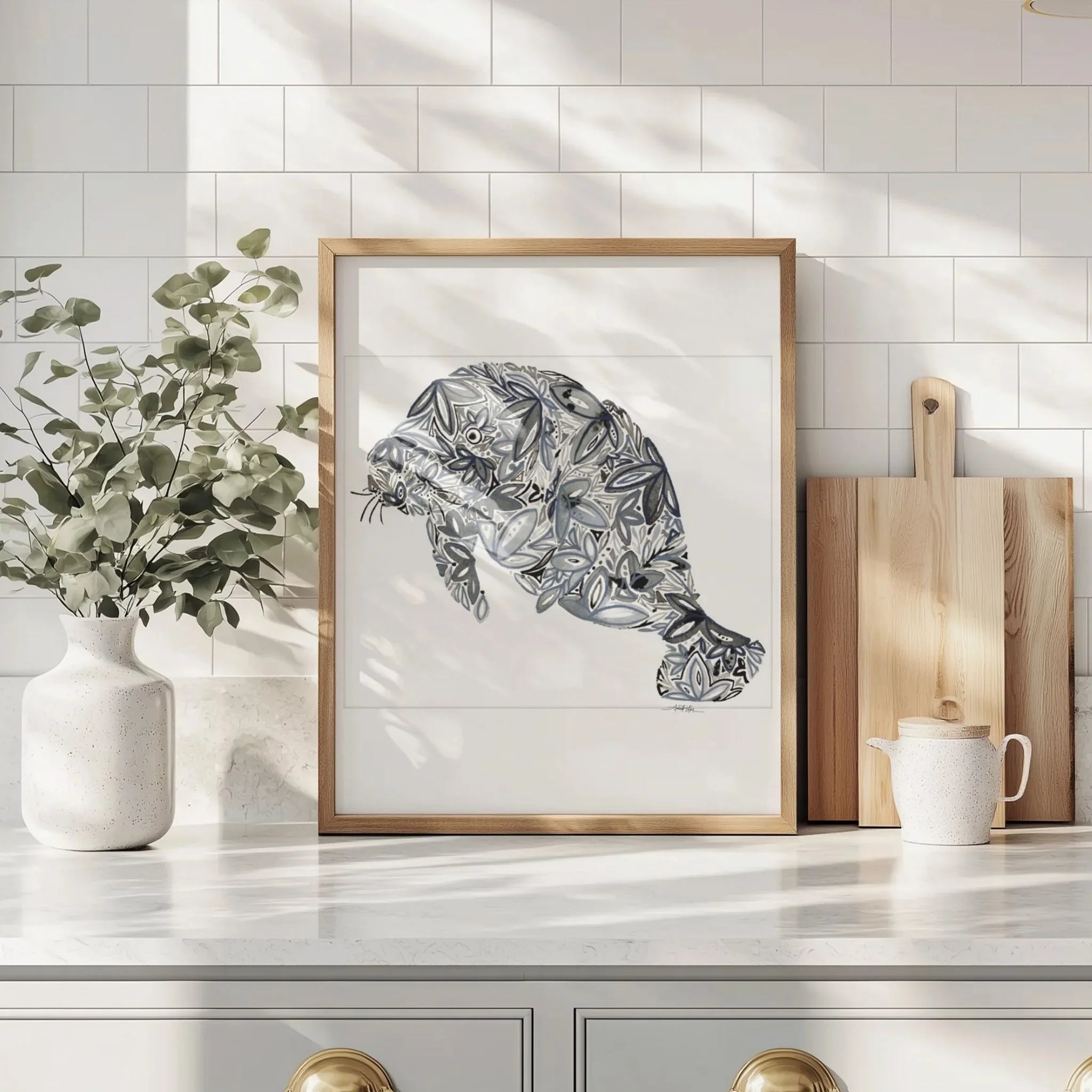 Framed manatee art print with floral patterns, displayed on kitchen counter beside greenery and cutting boards