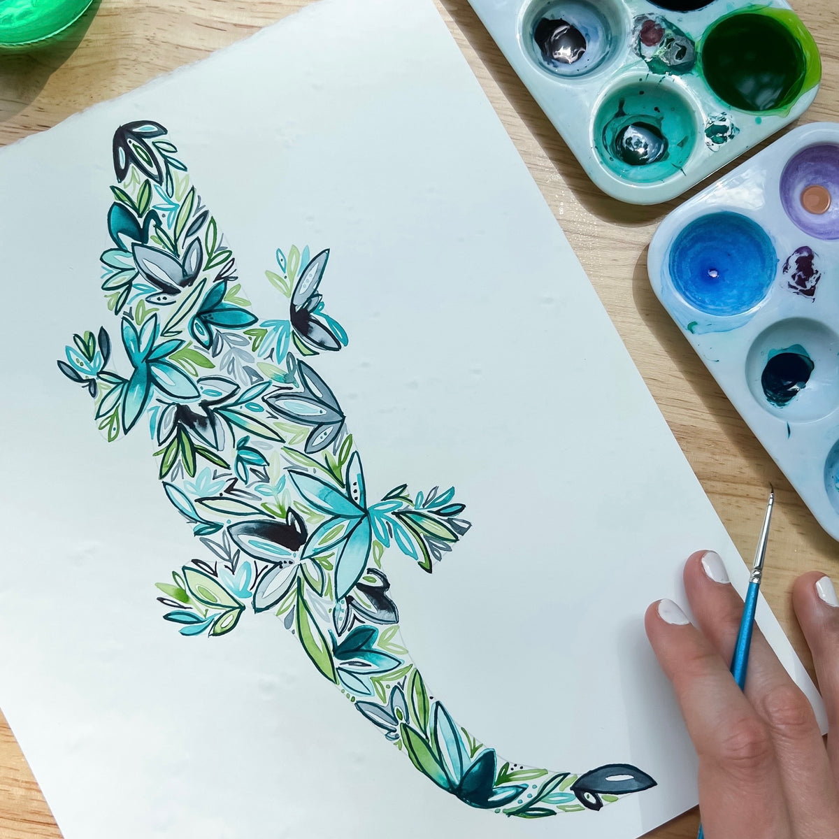 Hand-painted floral alligator art with watercolor palette and brush on wooden table